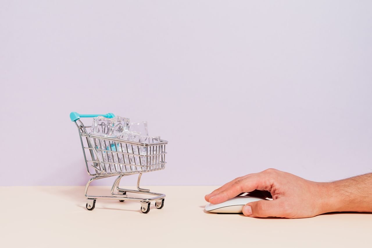 hero-img-02 Conceptual image of a hand on a mouse next to a miniature shopping cart filled with ice cubes, symbolizing online shopping.